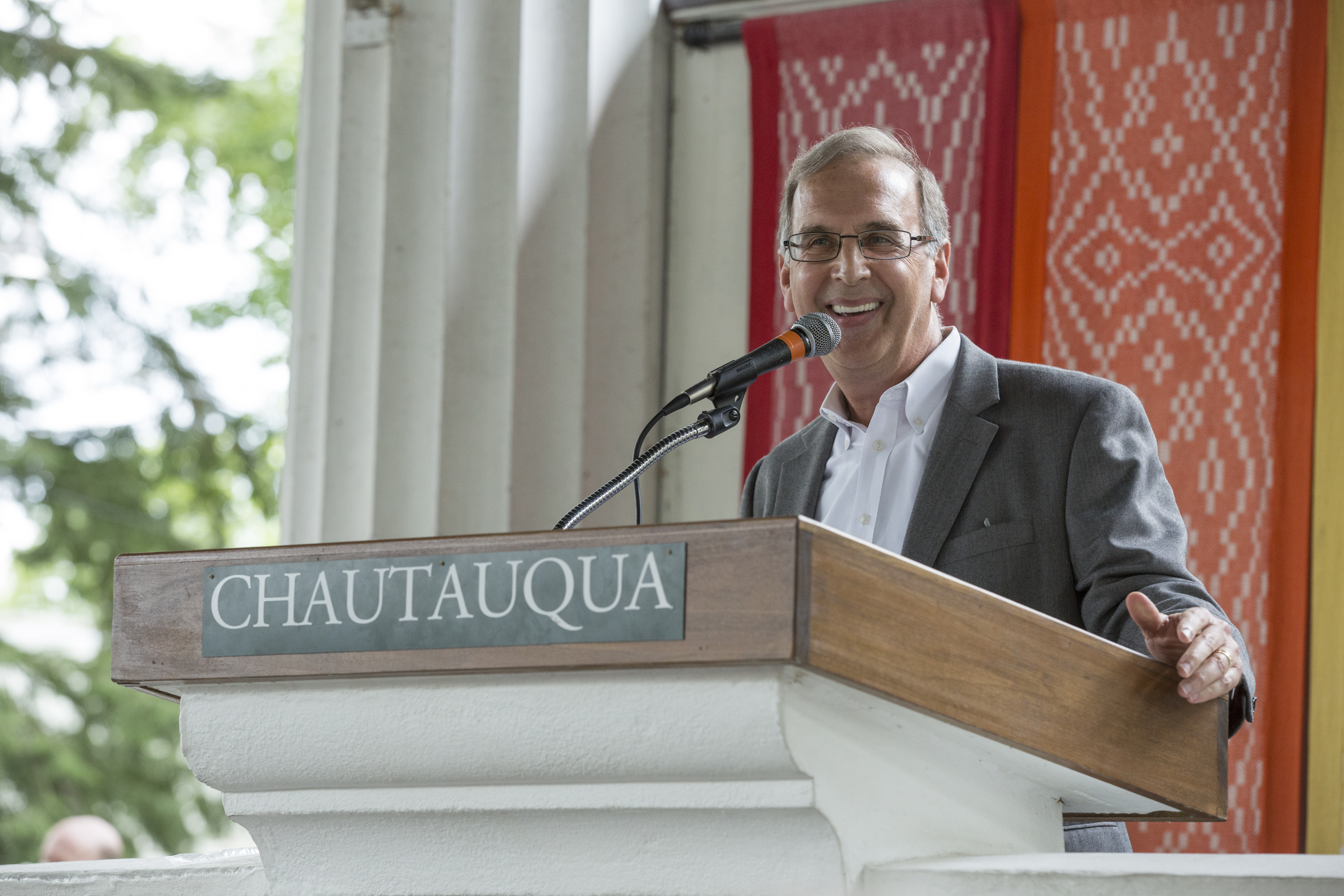 Rabbi Sid Schwarz smiling, standing at a podium with a sign on it reading "CHAUTAUQUA", on an outdoor stage with trees and a rainbow wall hanging behind him